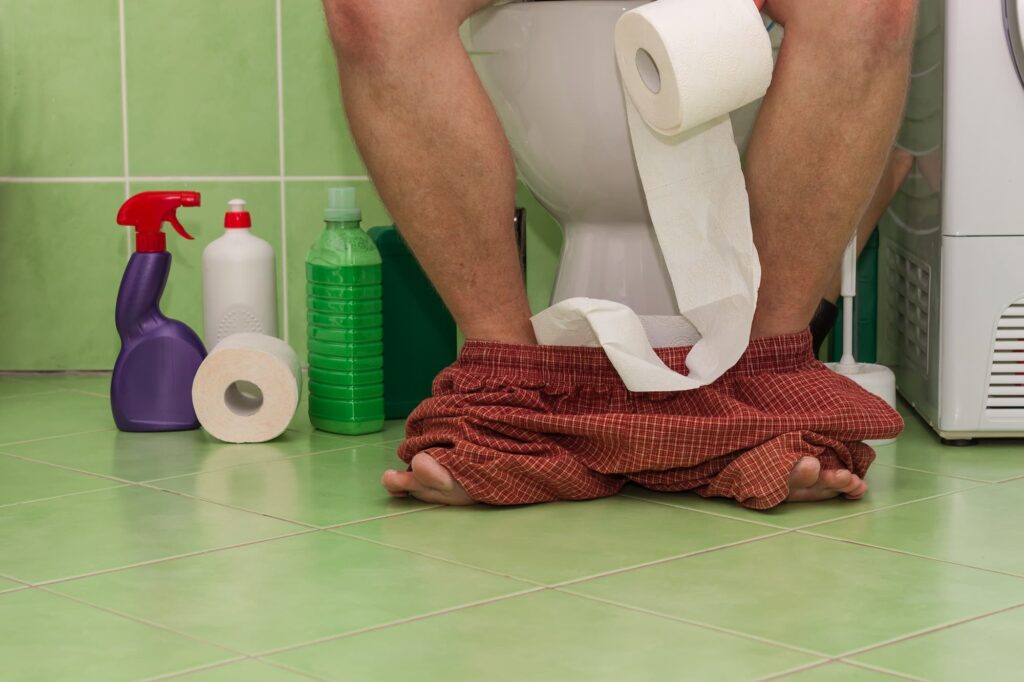 Piernas de un hombre sentado en el baño, sujetando un rollo de papel higiénico